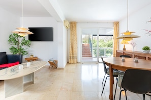A modern living room with a marble coffee table, a sofa, a TV mounted on the wall, and a dining area with a wooden table and chairs. - Casa Torremuelle II – Townhouse with private Pool & BBQ near Playa Bonita (Benalmádena)