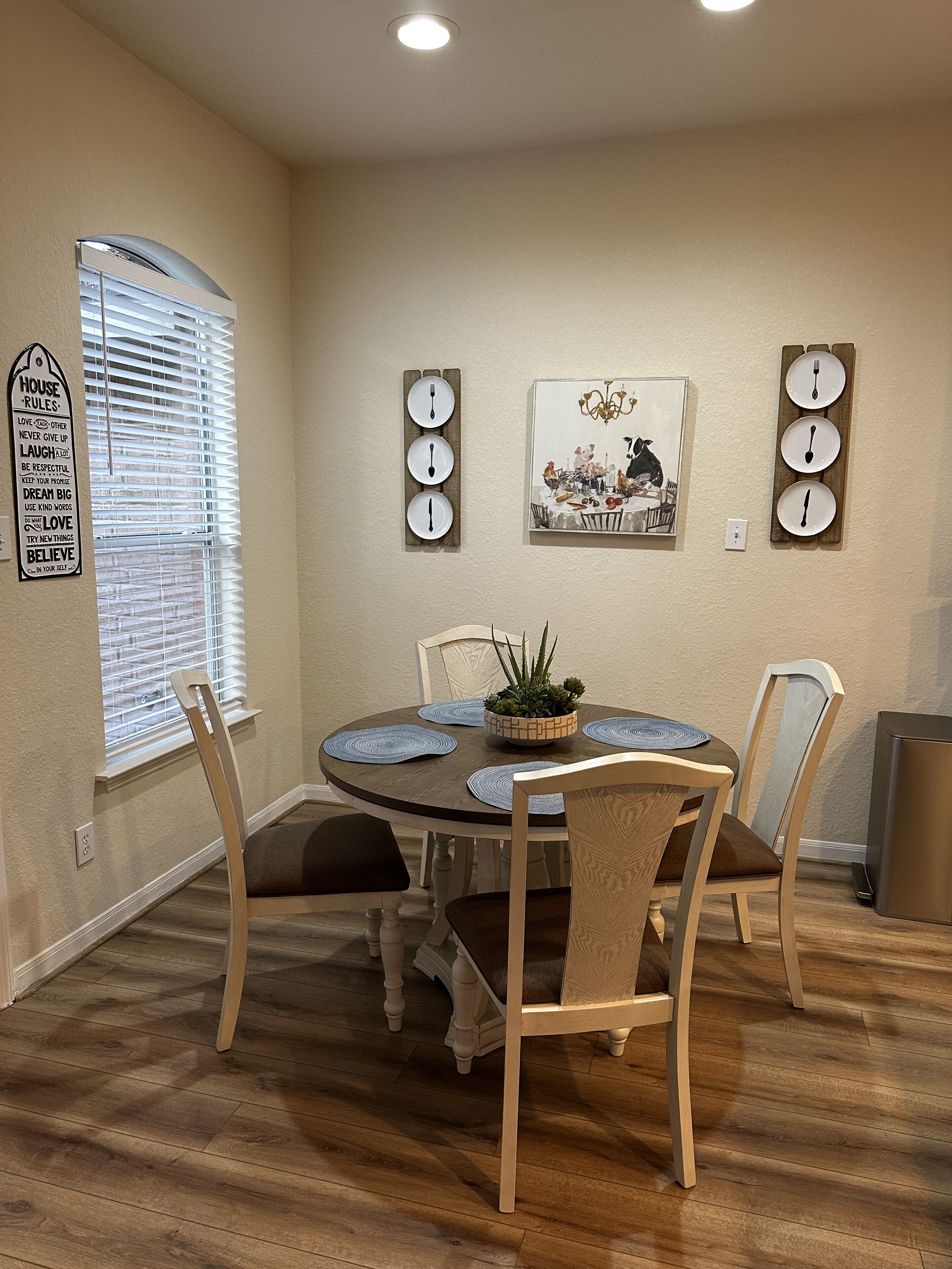 Uma sala de jantar com uma mesa redonda, quatro cadeiras, um vaso de planta, relógios de parede e um quadro emoldurado.