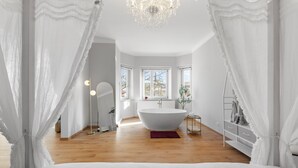 A modern bedroom with a large white bathtub, a chandelier, and sheer curtains.