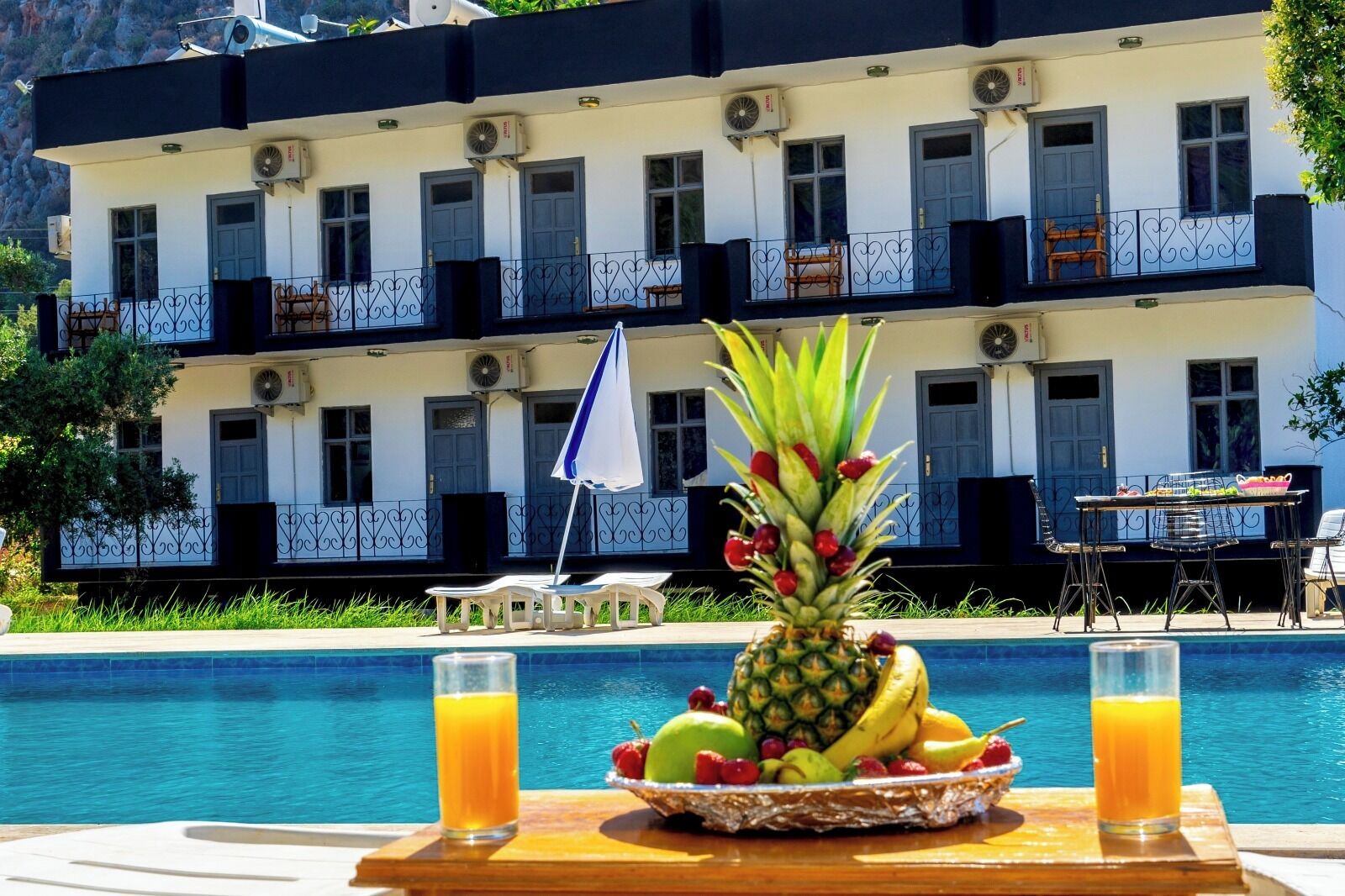 Desayuno junto a la piscina con una bandeja de frutas y dos vasos de jugo de naranja, una sombrilla blanca y una mesa con sillas.