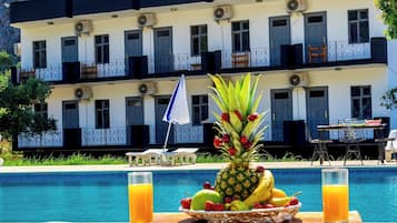 Desayuno junto a la piscina con una bandeja de frutas y dos vasos de jugo de naranja, una sombrilla blanca y una mesa con sillas.