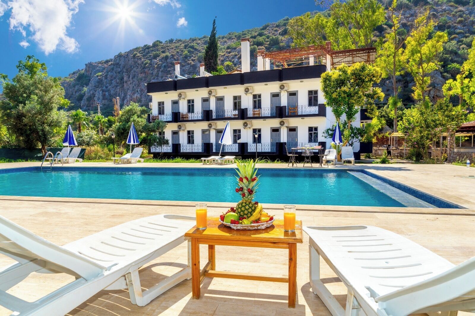 Uma área à beira da piscina com duas espreguiçadeiras, uma mesa de madeira com frutas e bebidas, e um edifício com varandas ao fundo.