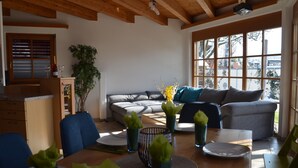 Ein gemütliches Wohnzimmer mit Holzbalkendecke, einem großen Fenster, einem grauen Sofa und einem Esstisch mit grünen Servietten.