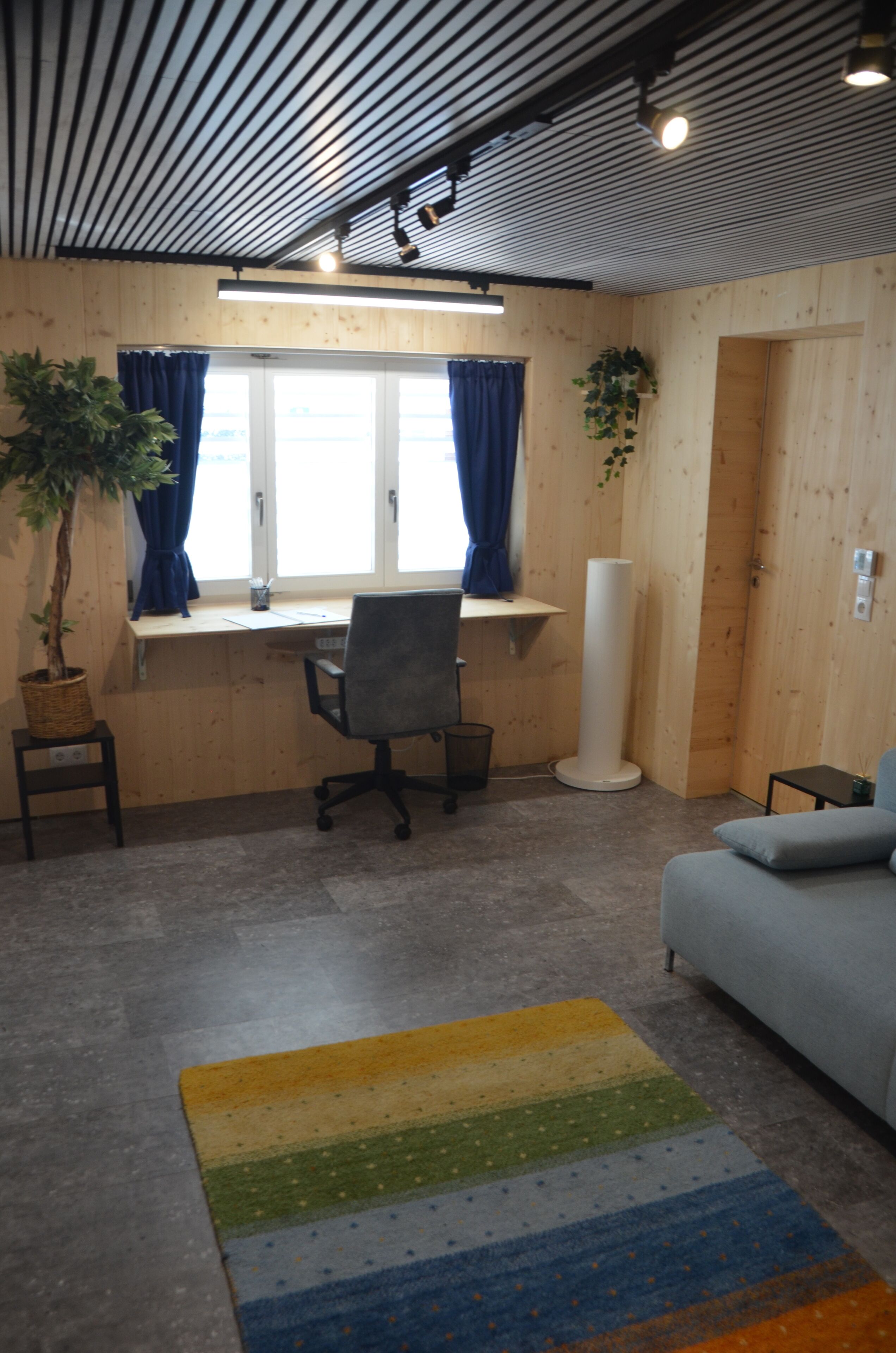 Ein Zimmer mit Holzvertäfelung, einem Schreibtisch mit Stuhl, einem Sofa, einer großen Topfpflanze und einer Stehlampe.