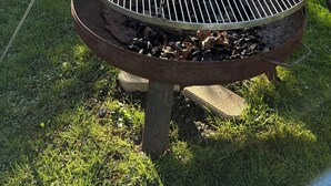 Ein runder, metallischer Grill mit Kettenstativ, auf einem Betonfundament in einem grünen Hinterhof platziert.