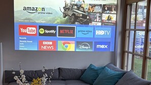 Ein modernes Wohnzimmer mit einer Eckcouch, einem Couchtisch mit Blumenvase und einem großen Bildschirm, der verschiedene Streaming-Dienste anzeigt.
