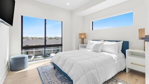 A modern bedroom with a large bed, two bedside tables, a TV, and a view of the city through large windows.