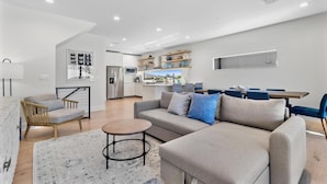 A modern living room with a sofa, armchair, and coffee table. There is a kitchen area with a refrigerator, microwave, and shelves.