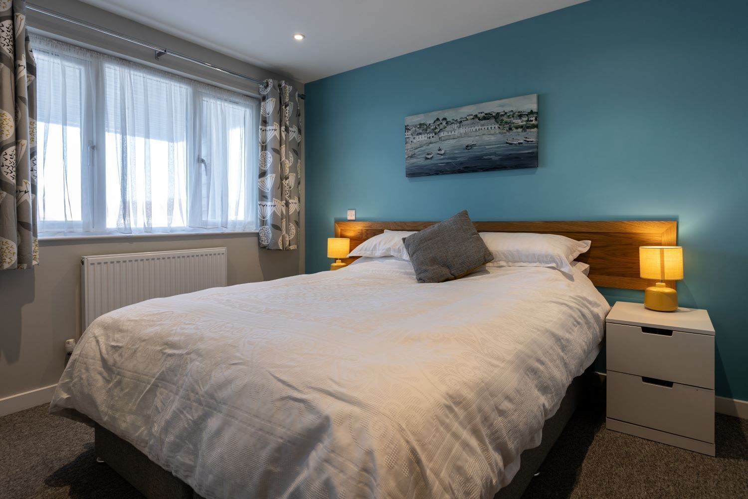 Een net opgemaakt bed met een houten hoofdbord, een nachtkastje met een lamp, en een schilderij van een kustlandschap aan de muur.