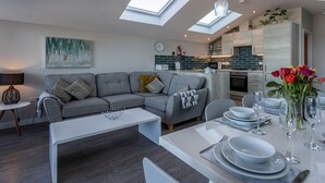 A modern living room with a grey sofa, a white coffee table, and a dining area set for a meal. - 3 Bed pet friendly Beach House with sea views (Westward Ho!)