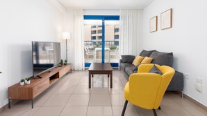 En moderne stue med et fladskærms-tv, en gul lænestol, en grå sofa og en balkon med blåt gelænder.