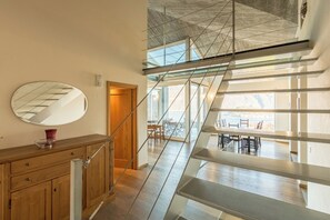 Um interior moderno com um armário de madeira, um espelho redondo e uma escada que leva a um andar superior com guarda-corpo de vidro e vista para o exterior.