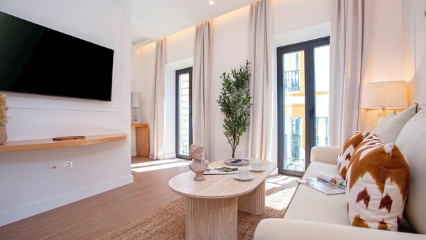 Een moderne woonkamer met een flatscreen televisie, een ronde salontafel, een bank met opvallende kussens en een groot raam met gordijnen.