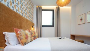 Een net opgemaakt bed met witte en gemêleerde beddengoed, een houten hoofdbord en een nachtkastje met een lamp.
