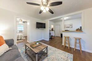 Una sala moderna con ventilador de techo, televisor, comedor con mesa y sillas, y zona de cocina con taburetes.