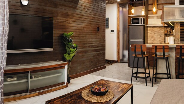 A modern living room with a flat-screen TV, wooden wall paneling, a ceiling fan, and a dining area with bar stools. - Chic 1BR Retreat: Steps to Rasfannu Beach & Malé’s Best (Malé)