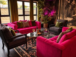 A room with a vibrant pink sofa, a dark armchair, and a glass coffee table. There is a large window with curtains, a potted plant, and a decorative wall. - Romantic  Couples Getaway • W/Fireplace &Private Patio • Walk Everywhere (Saugatuck)