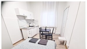 A compact room with a bed, a small kitchenette, a dining table with chairs, and a window with curtains.