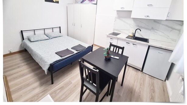 A compact bedroom with a bed, bedside tables, and a wardrobe. Adjacent is a kitchenette with a sink, stove, and cabinets.