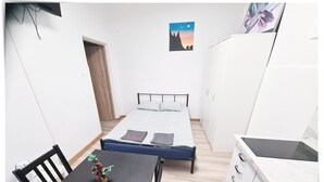 A small, well-lit room with a bed, a table with a potted plant, a white wardrobe, and a small kitchenette.