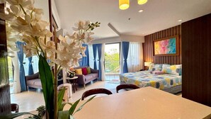 Een moderne hotelkamer met een bank, een bed met kleurrijk beddengoed, een eettafel en een groot raam met gordijnen.