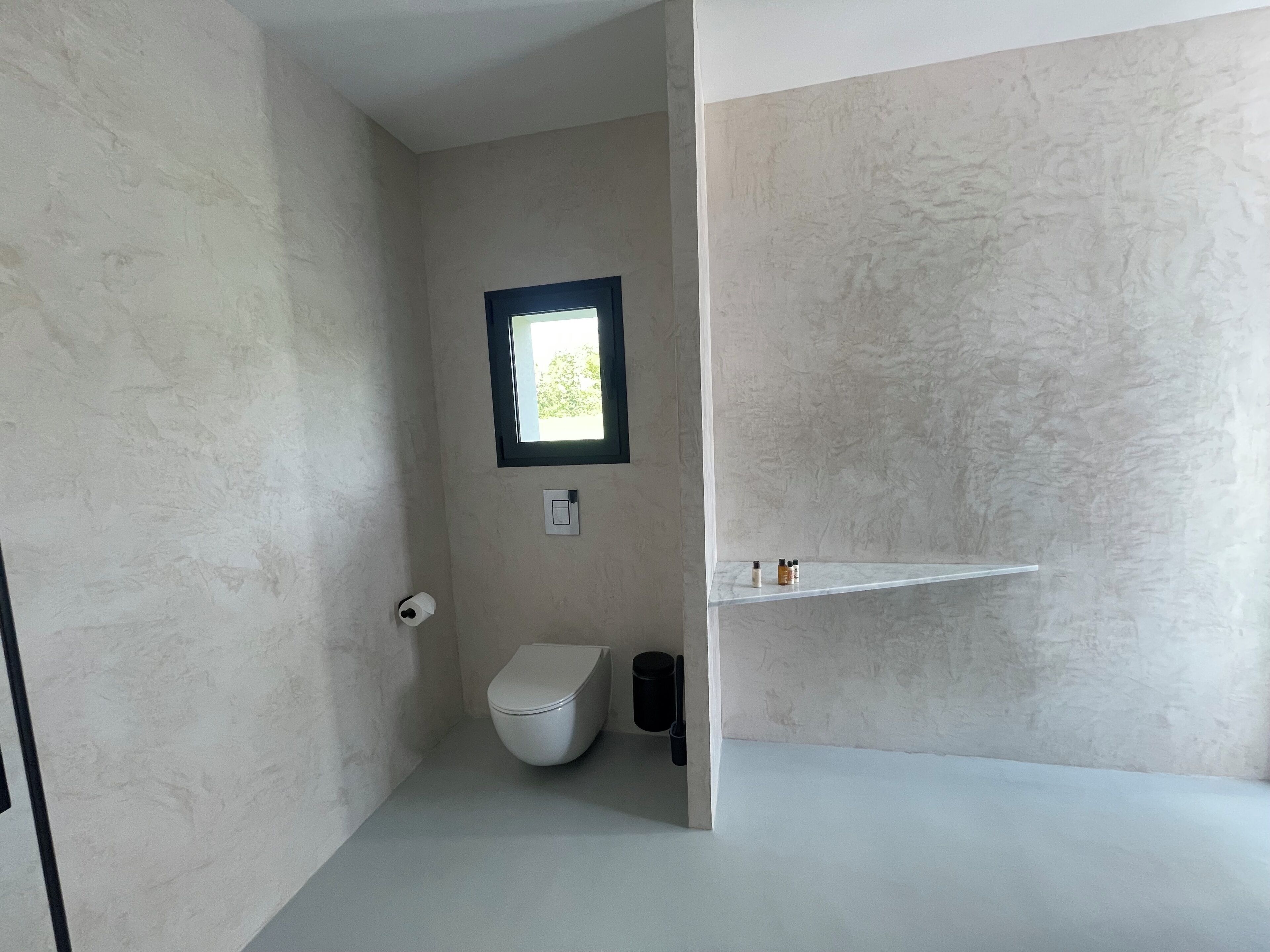 A bathroom with a toilet, a small window, and a shelf with toiletries.