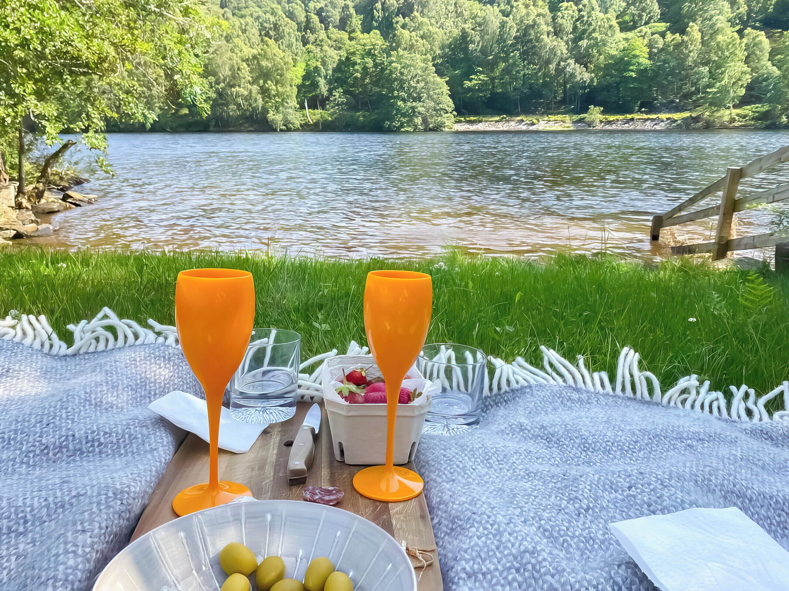 Une table en bois dressée au bord d’une rivière pour un pique-nique, avec une couverture, deux verres orange, un bol d’olives et un couteau.