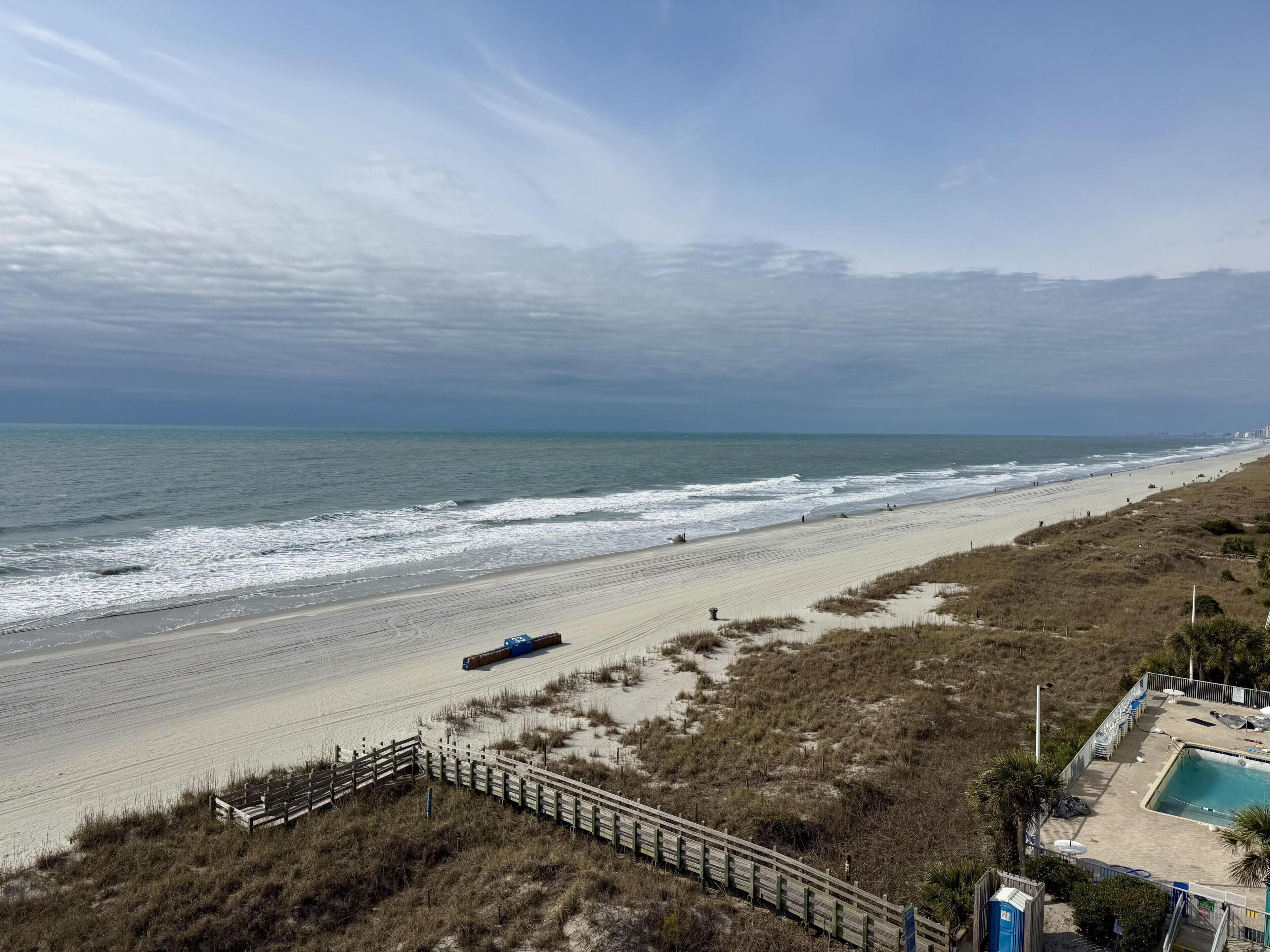 A beach view with a wooden walkway, a swimming pool, and a distant city skyline.