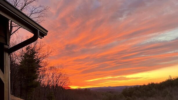 A sunset view from a deck with a railing, overlooking a forested area and a distant mountain range. - Spacious mountain home 3mi south of Blowing Rock, NC, views, creek, 56 acres. (Lenoir)
