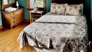 A bedroom with a wooden dresser, a bed with floral bedding, a ceiling fan, a window with a view, and a framed picture on the wall. - Spacious mountain home 3mi south of Blowing Rock, NC, views, creek, 56 acres. (Lenoir)