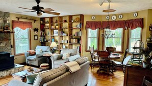 A cozy living room with a fireplace, a ceiling fan, and a dining area with a table and chairs. - Spacious mountain home 3mi south of Blowing Rock, NC, views, creek, 56 acres. (Lenoir)