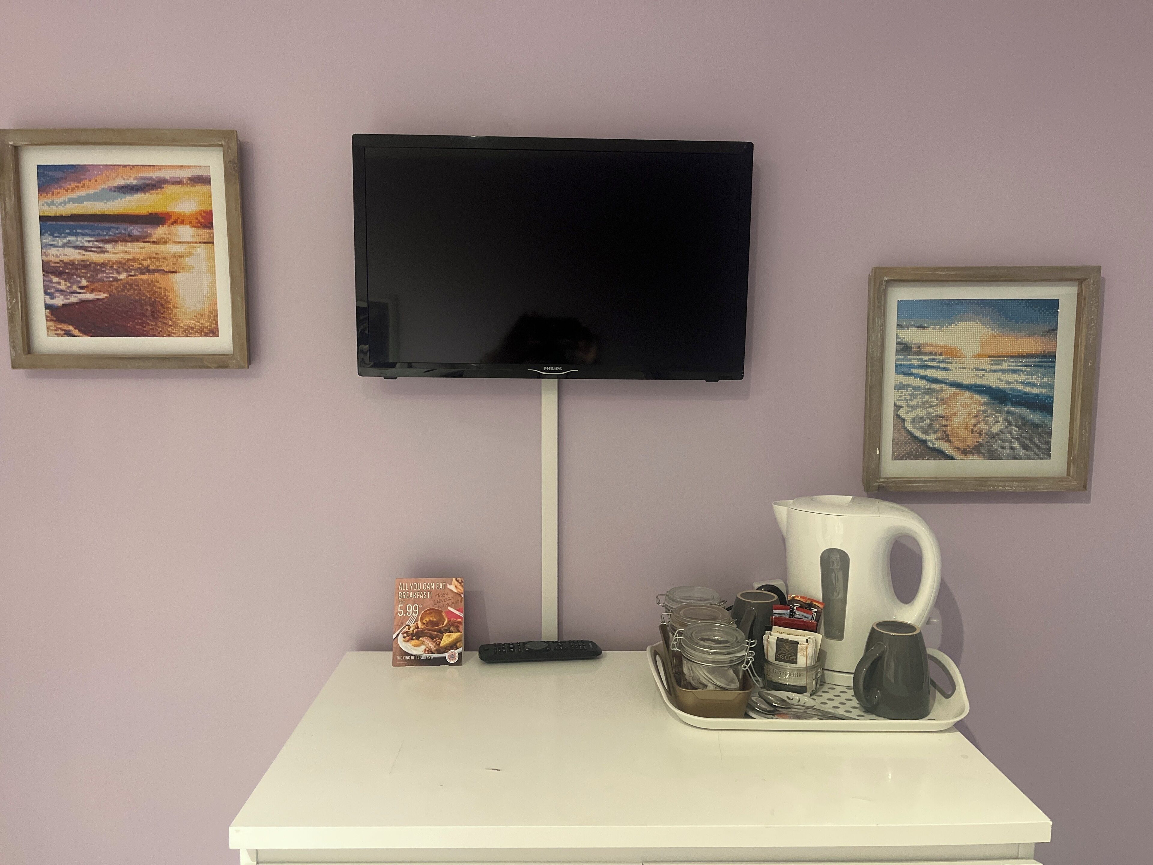 A flat-screen TV mounted on a wall, a white table with a tray holding a coffee maker, cups, and a remote control.