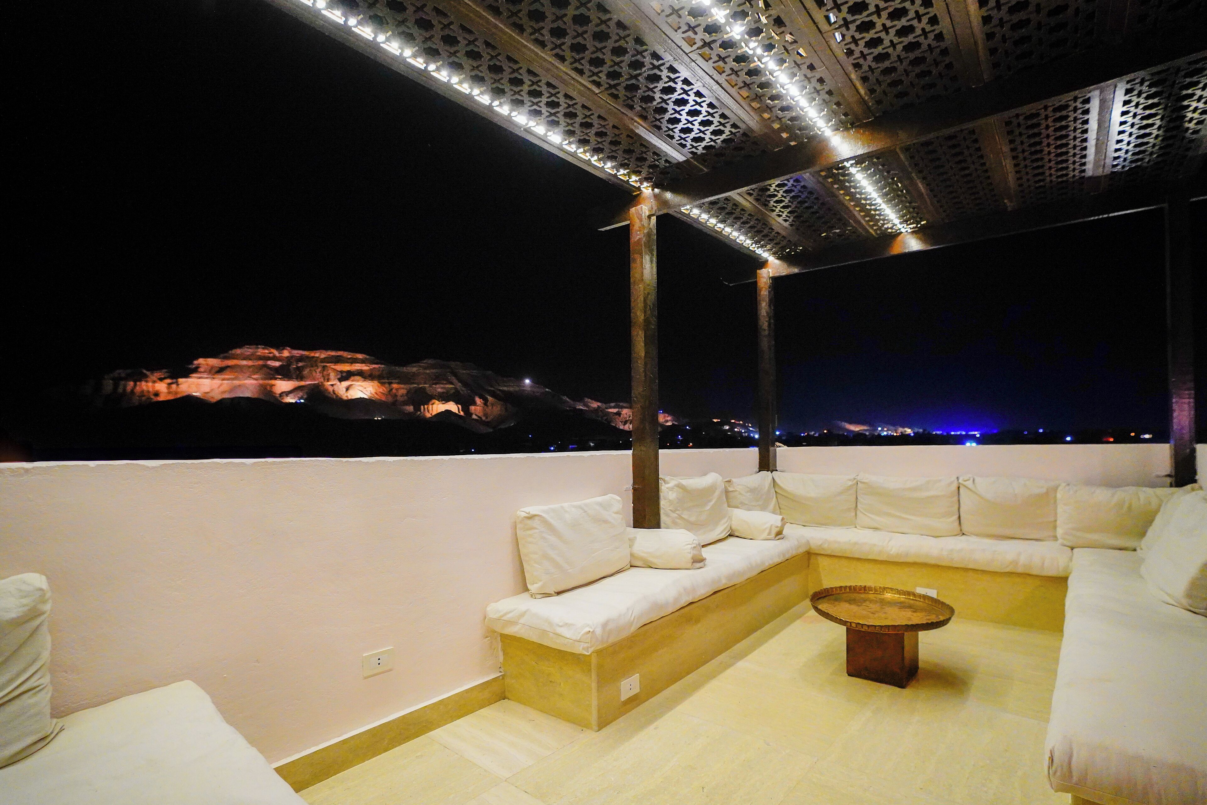 A rooftop terrace with a white sofa, a round coffee table, and a view of a cityscape at night.