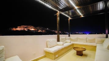 A rooftop terrace with a white sofa, a round coffee table, and a view of a cityscape at night.