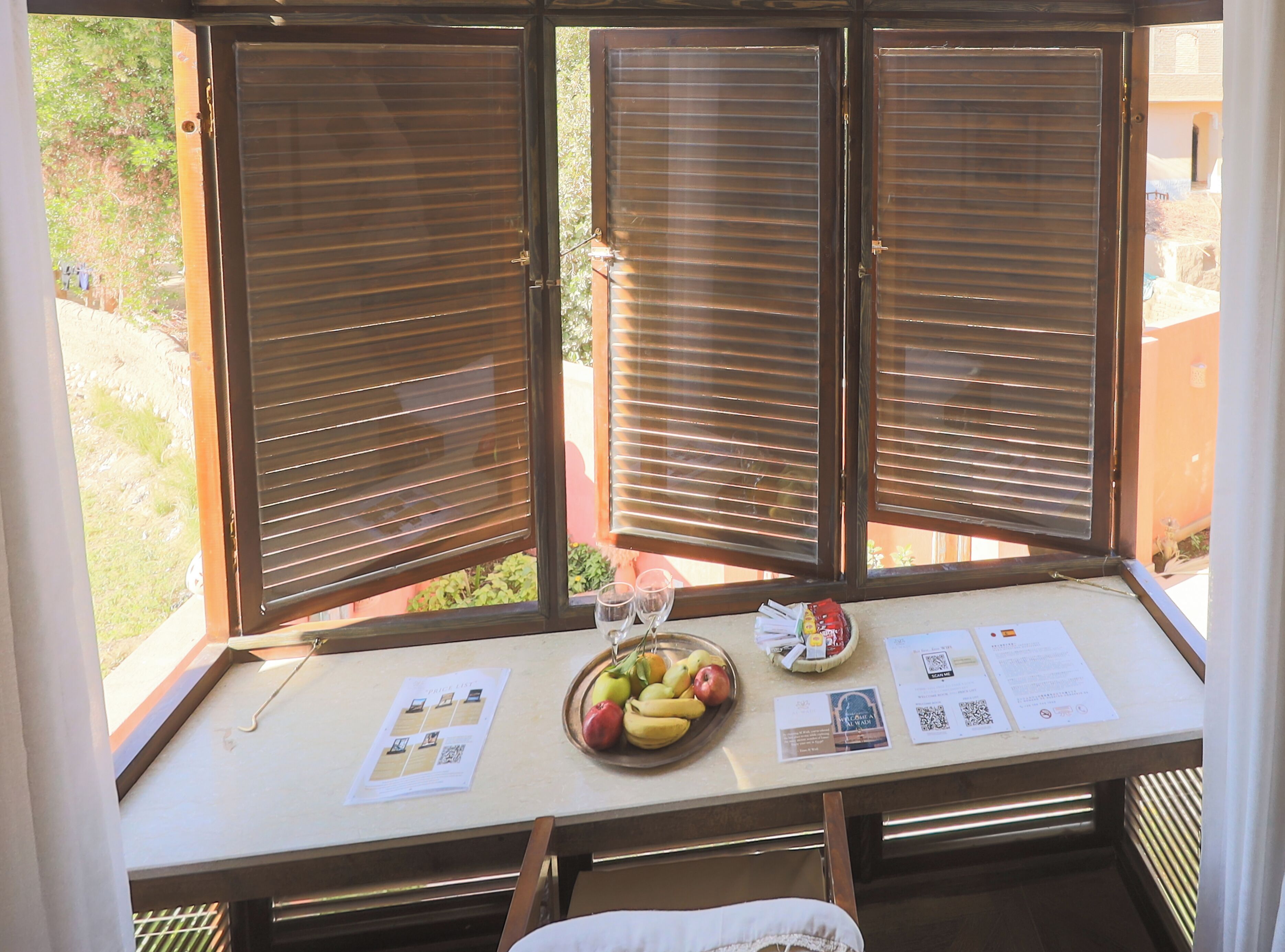 A window with closed blinds, a table with a fruit bowl, a glass, and some papers.