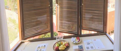 A window with closed blinds, a table with a fruit bowl, a glass, and some papers.