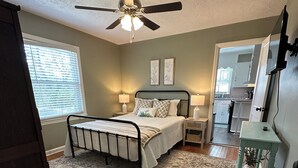 Une chambre à coucher comprenant un lit, des tables de chevet, un ventilateur de plafond, un tapis et une vue sur la cuisine adjacente.