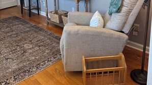 Un salon avec un fauteuil gris, une table d’appoint en bois, un téléviseur à écran plat, un tapis à motifs et un petit berceau en bois.