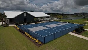 A tennis court with a fenced area, a covered seating area, and a building with a white roof. -  RV Lot 67 in Grand Riviera Resort in Foley, AL. Daily and long term available (Foley)