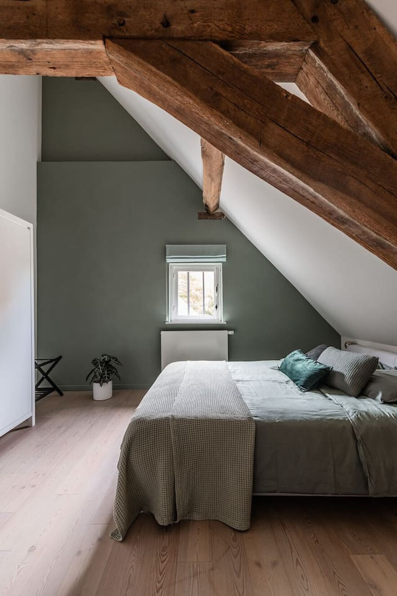 Une chambre avec un plafond en pente, une fenêtre, un lit avec des oreillers, un radiateur et une plante en pot.
