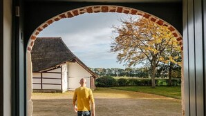 A man walking through a covered entrance with a brick archway and a view of a building with a thatched roof.