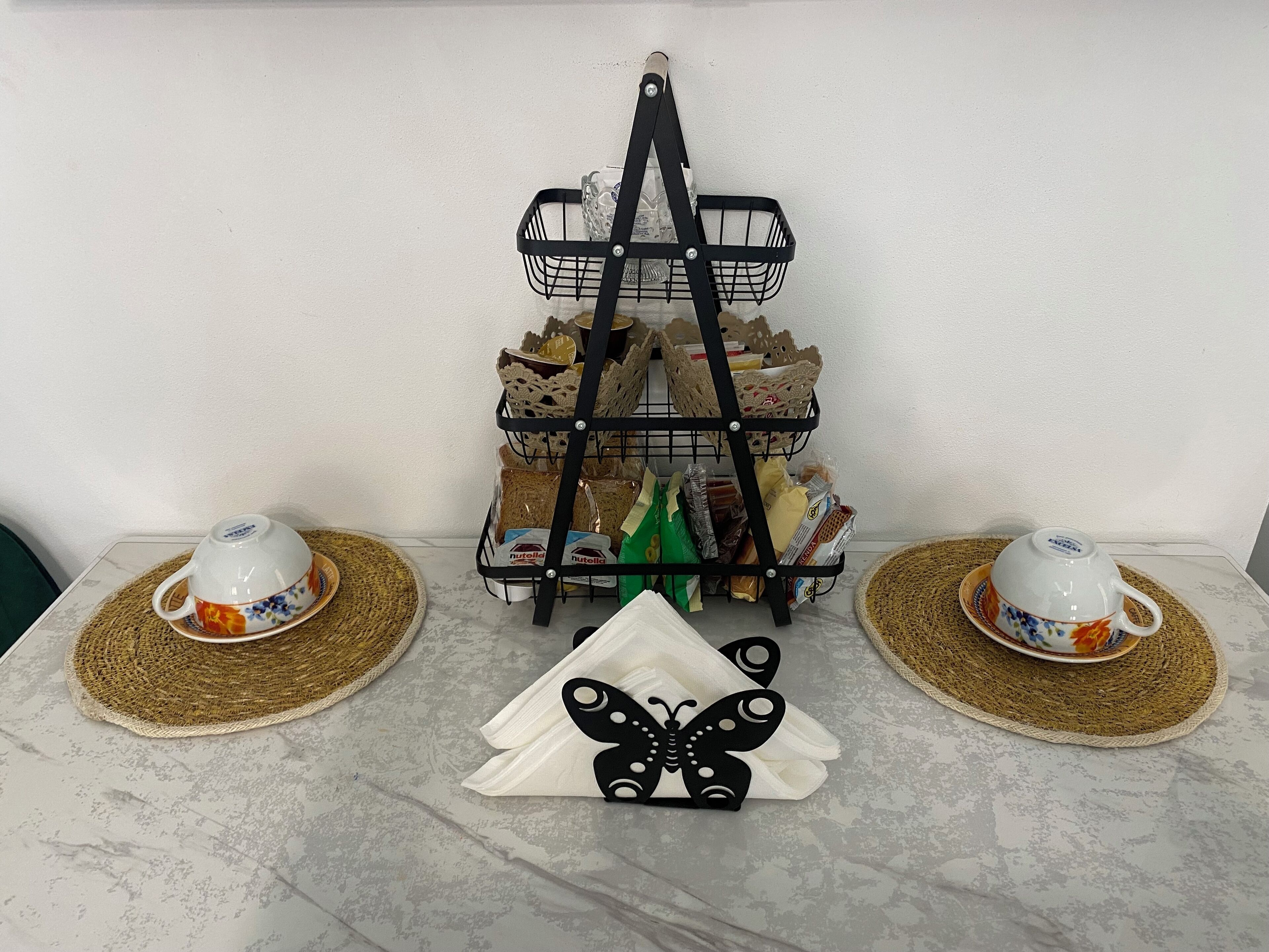 Une étagère en métal noir à trois niveaux, sur laquelle sont disposés divers objets, deux tasses à thé blanches ornées de motifs floraux et un porte-serviettes noir en forme de papillon, le tout posé sur une table en marbre.
