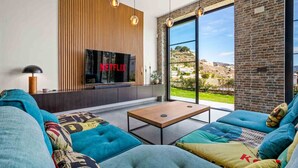 A modern living room with a large TV screen displaying 'NETFLIX', a sofa set, a coffee table, and a view of a hilly landscape through large windows. - Casa Hurlavente | Luxury Villa on Punta de Mona | 4 En-suite Bedrooms | Heated Private Pool | Panoramic Sea Views | Roof Terrace (La Herradura)