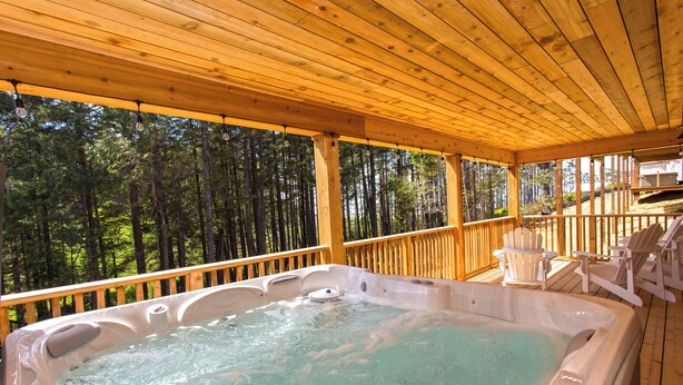 Une terrasse en bois avec un jacuzzi, entourée de chaises longues blanches et offrant une vue sur une forêt.