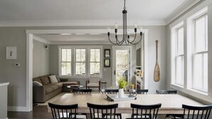 Un salon spacieux avec une grande table à manger, un lustre et une vue sur l’extérieur grâce à de grandes fenêtres.