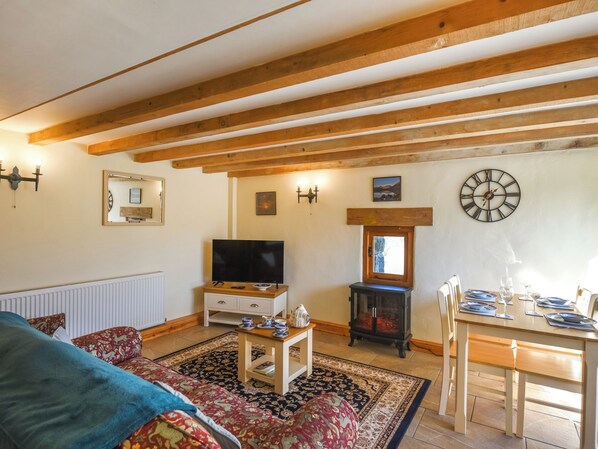 A cozy living room with a TV, wooden beams, a fireplace, a dining table with chairs, and a patterned rug.