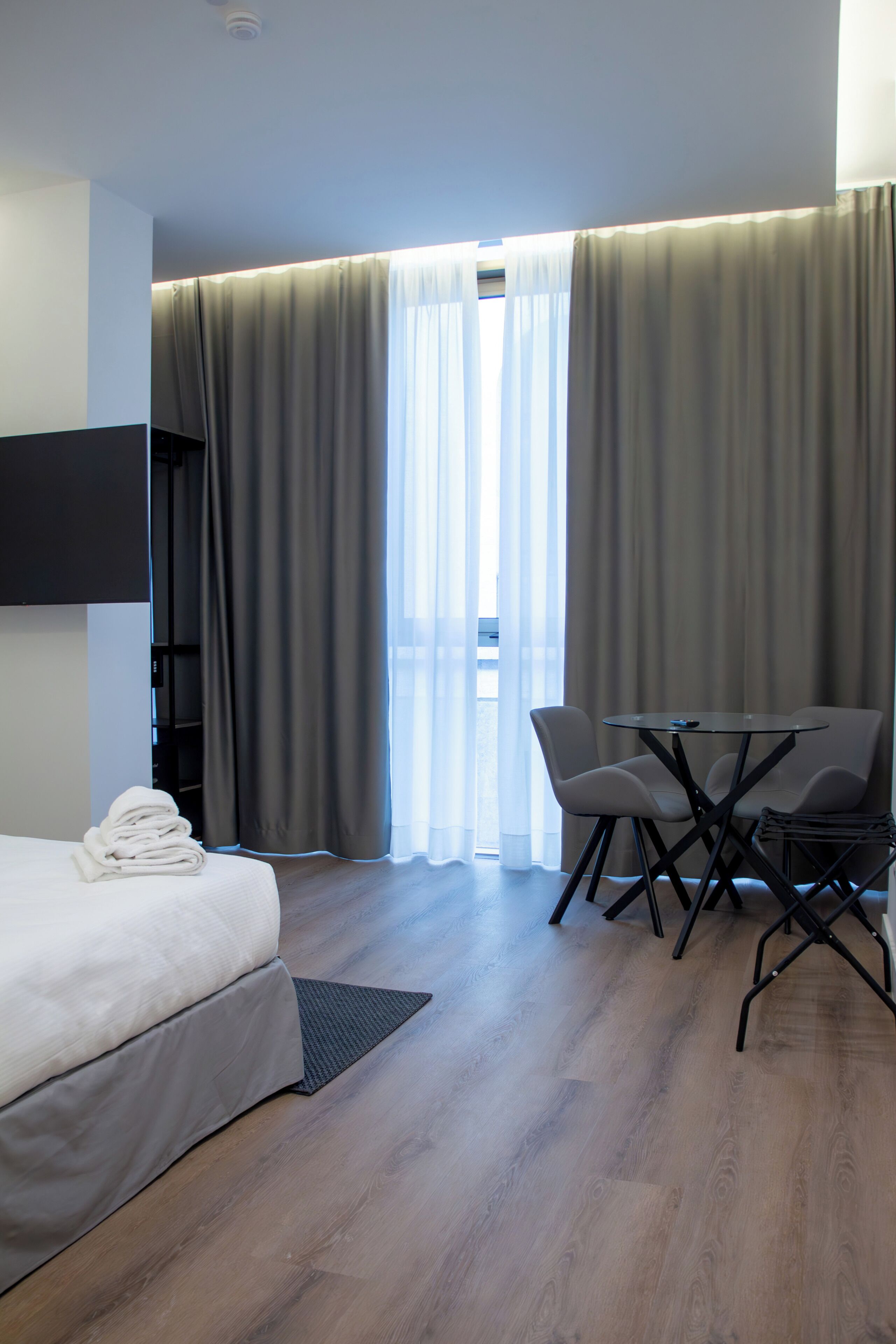 Quarto de hotel com cama, secretária com cadeiras, televisão e janela com cortinas.