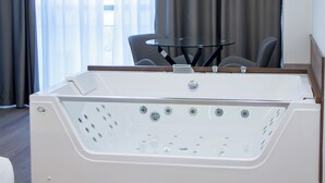 Une salle de bain moderne avec une grande baignoire blanche, une table en verre et une vue sur l’extérieur à travers des rideaux transparents.