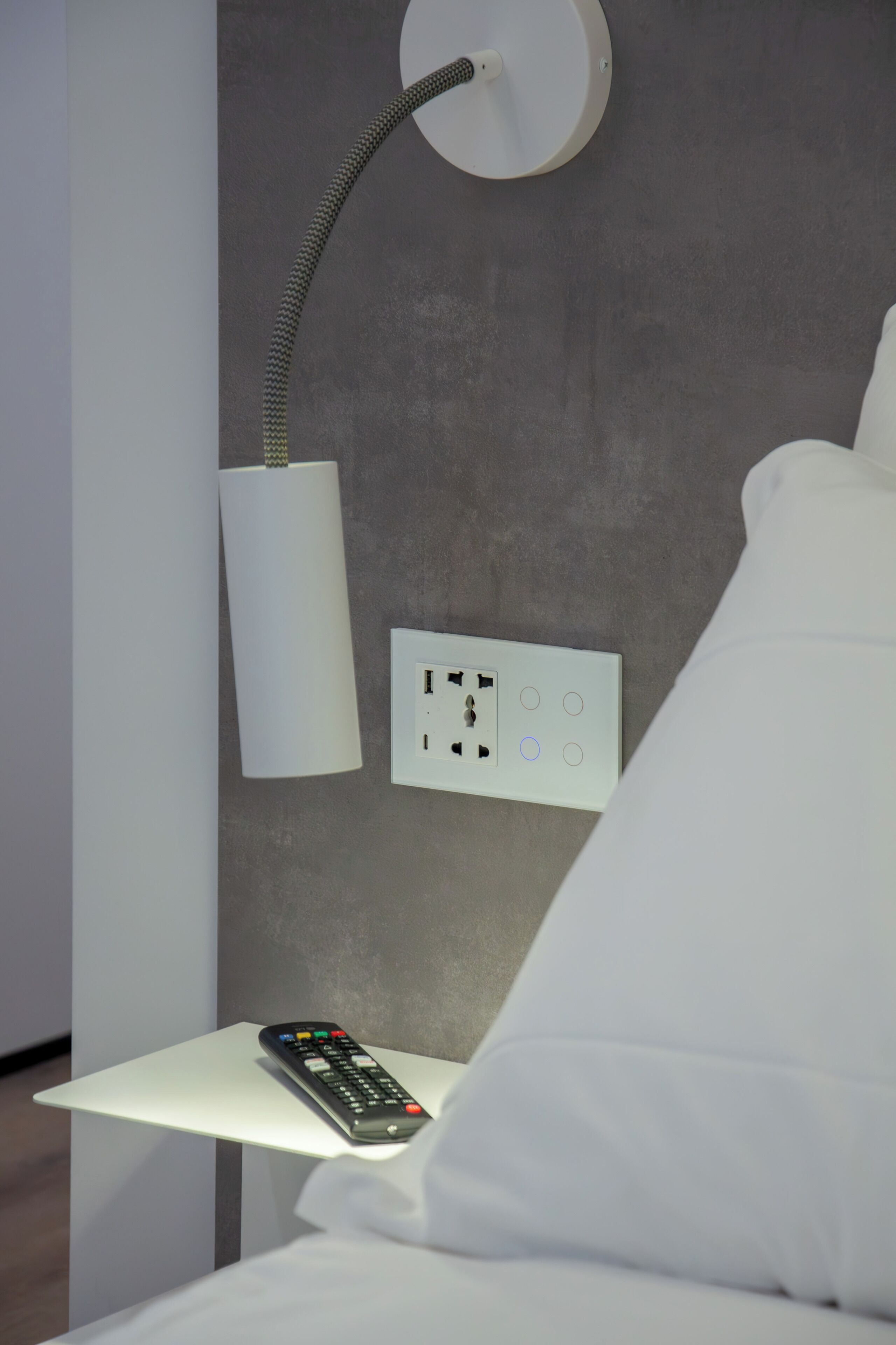 Una cama bien hecha con ropa blanca, un control remoto sobre una mesita de noche blanca, una lámpara de pared y un enchufe.