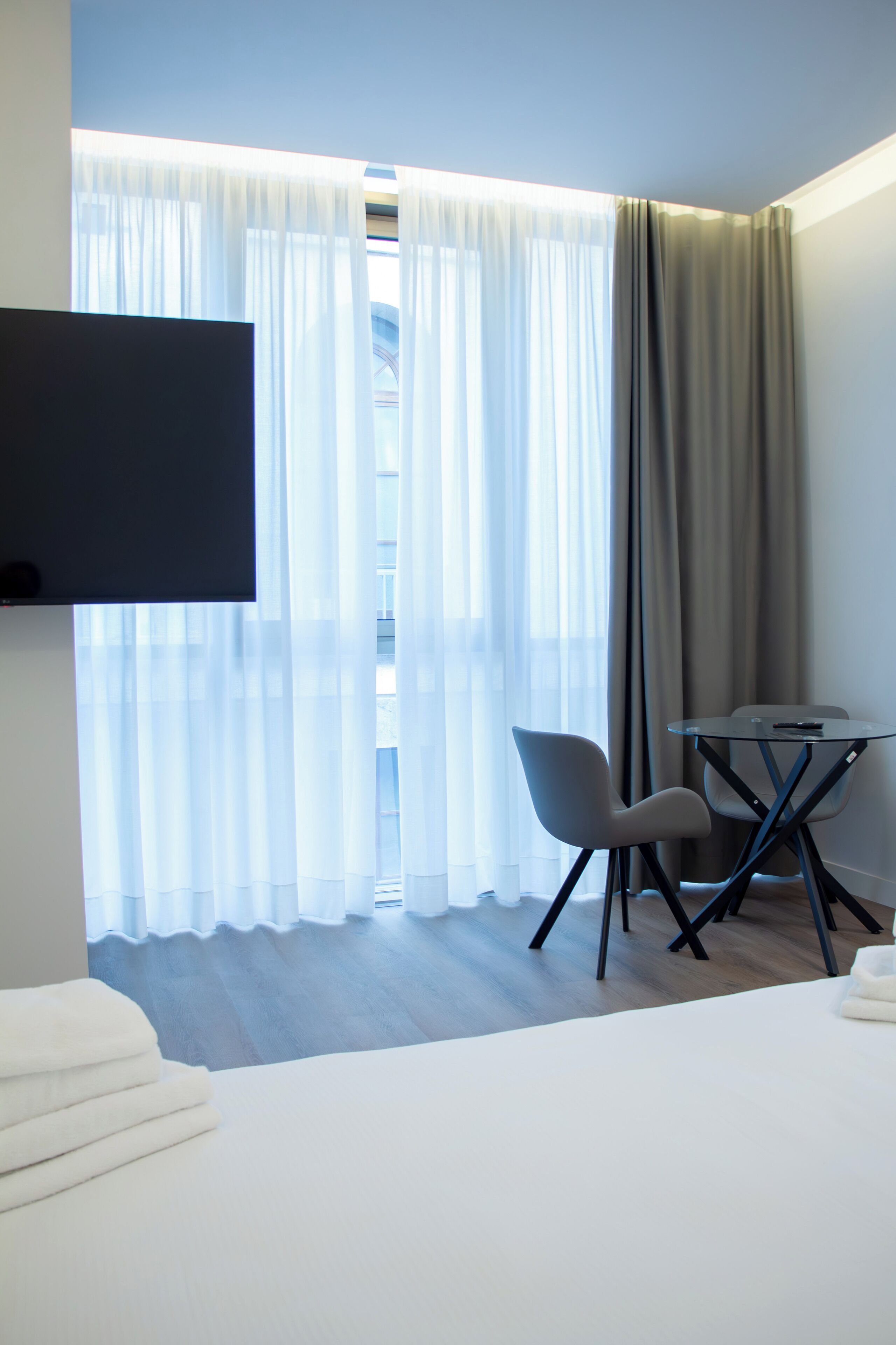 A modern hotel room with a bed, a flat-screen TV mounted on the wall, a small table with two chairs, and a window with sheer curtains.